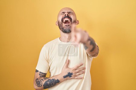 Foto de Hombre hispano con tatuajes de pie sobre fondo amarillo riéndose de ti, señalando con el dedo a la cámara con la mano sobre el cuerpo, expresión de vergüenza - Imagen libre de derechos