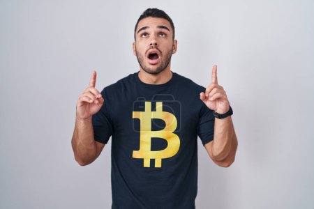 Téléchargez les photos : Jeune homme hispanique portant Bitcoin t-shirt étonné et surpris de regarder vers le haut et pointant avec les doigts et les bras levés. - en image libre de droit