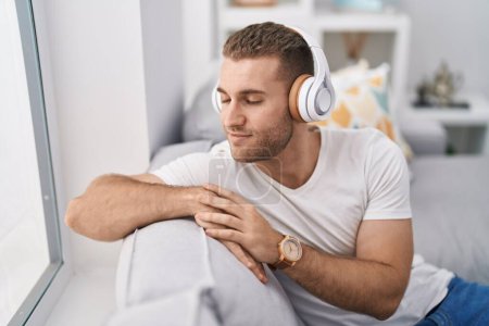 Téléchargez les photos : Jeune homme caucasien écoutant de la musique assis sur le canapé à la maison - en image libre de droit