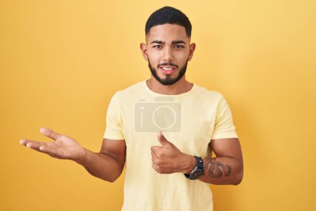 Téléchargez les photos : Young hispanic man standing over yellow background showing palm hand and doing ok gesture with thumbs up, smiling happy and cheerful - en image libre de droit