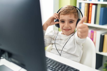 Téléchargez les photos : Adorable élève hispanique garçon utilisant ordinateur et écouteurs faisant geste pouce vers le haut à la salle de classe - en image libre de droit