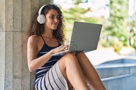 Photo for Young beautiful hispanic woman listening to music sitting on bench at street - Royalty Free Image
