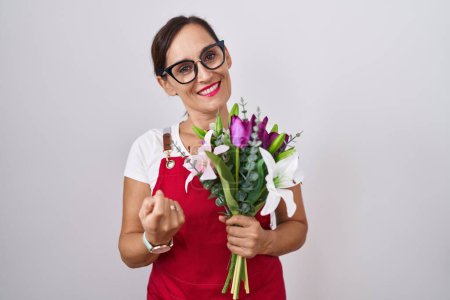 Téléchargez les photos : Femme brune du moyen âge portant un tablier travaillant dans un magasin de fleuristes tenant un bouquet, venez ici geste avec la main invitant à l'accueil heureux et souriant - en image libre de droit
