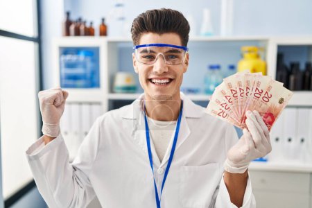 Photo for Young hispanic man working at scientist laboratory holding shekels screaming proud, celebrating victory and success very excited with raised arm - Royalty Free Image