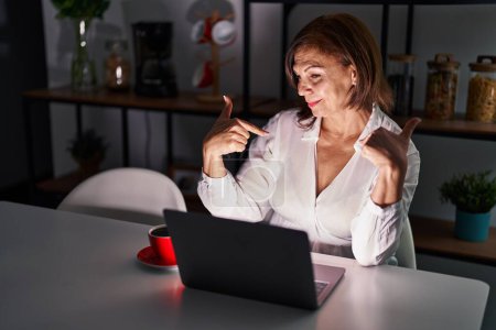 Foto de Mujer hispana de mediana edad que usa el portátil en casa por la noche y se ve segura de sí misma con sonrisa en la cara, señalándose con los dedos orgullosos y felices. - Imagen libre de derechos