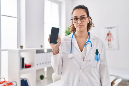 Photo for Young hispanic doctor woman holding smartphone showing screen thinking attitude and sober expression looking self confident - Royalty Free Image
