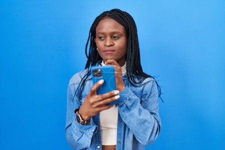 Photo for African woman with braids using smartphone typing message serious face thinking about question with hand on chin, thoughtful about confusing idea - Royalty Free Image