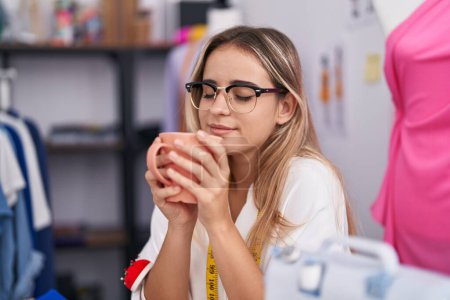 Téléchargez les photos : Jeune femme blonde tailleur souriant boire du café en toute confiance au magasin de vêtements - en image libre de droit