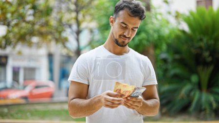 Téléchargez les photos : Jeune homme hispanique souriant confiant comptant pesos mexicains au parc - en image libre de droit