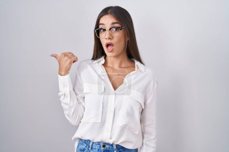 Photo for Young brunette woman wearing glasses surprised pointing with hand finger to the side, open mouth amazed expression. - Royalty Free Image