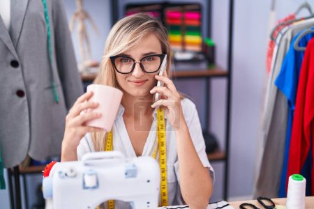Téléchargez les photos : Young blonde woman tailor talking on smartphone drinking coffee at tailor shop - en image libre de droit