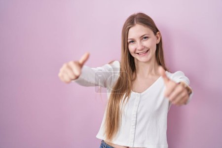 Foto de Joven mujer caucásica de pie sobre fondo rosa aprobando hacer gesto positivo con la mano, pulgares hacia arriba sonriendo y feliz por el éxito. gesto ganador. - Imagen libre de derechos