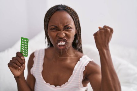 Foto de Mujer afroamericana sosteniendo píldoras anticonceptivas molesto y frustrado gritando con ira, gritando loco de ira y la mano levantada - Imagen libre de derechos