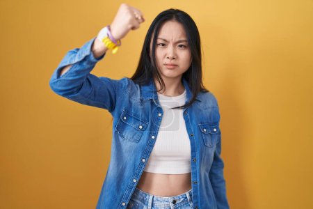 Téléchargez les photos : Jeune femme asiatique debout sur fond jaune en colère et fou levant le poing frustré et furieux tout en criant de colère. rage et concept agressif. - en image libre de droit