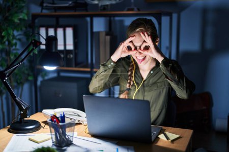 Photo for Young blonde woman working at the office at night doing ok gesture like binoculars sticking tongue out, eyes looking through fingers. crazy expression. - Royalty Free Image