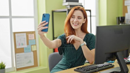 Foto de Joven pelirroja trabajadora de negocios utilizando la computadora hacer selfie por teléfono inteligente en la oficina - Imagen libre de derechos