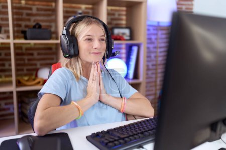 Foto de Joven mujer caucásica jugando videojuegos usando auriculares rezando con las manos juntas pidiendo perdón sonriendo confiado. - Imagen libre de derechos