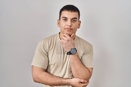 Téléchargez les photos : Jeune homme arabe portant t-shirt décontracté regardant avec confiance la caméra souriant avec les bras croisés et la main levée sur le menton. penser positivement. - en image libre de droit