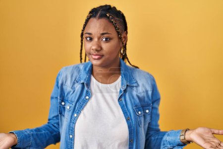 Photo for African american woman with braids standing over yellow background clueless and confused with open arms, no idea concept. - Royalty Free Image
