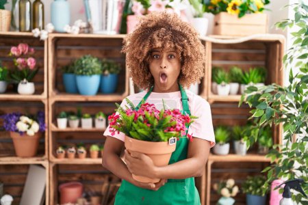 Photo for Young hispanic woman with curly hair working at florist shop holding plant scared and amazed with open mouth for surprise, disbelief face - Royalty Free Image