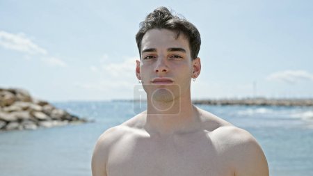 Téléchargez les photos : Jeune homme hispanique debout touristique avec expression détendue à la plage - en image libre de droit