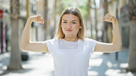 Téléchargez les photos : Jeune femme blonde souriante confiante faisant un geste fort avec les bras à la rue - en image libre de droit