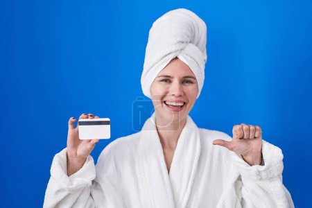 Téléchargez les photos : Blonde femme caucasienne portant peignoir tenant carte de crédit pointant pouce vers le côté souriant heureux avec bouche ouverte - en image libre de droit