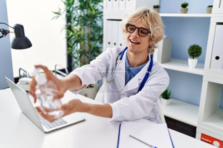 Téléchargez les photos : Jeune homme blond médecin souriant confiant à l'aide de gel désinfectant mains à la clinique - en image libre de droit