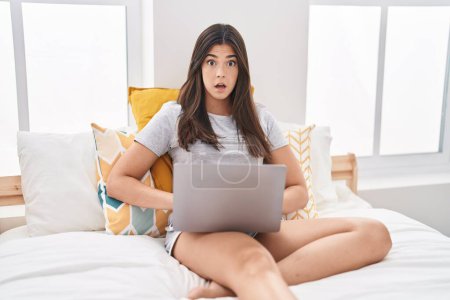 Photo for Hispanic woman using computer laptop on the bed scared and amazed with open mouth for surprise, disbelief face - Royalty Free Image