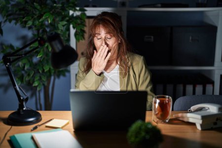 Téléchargez les photos : Moyen Age femme hispanique travaillant à l'aide d'un ordinateur portable la nuit ennuyé bâillement fatigué couvrant la bouche avec la main. agitation et somnolence. - en image libre de droit