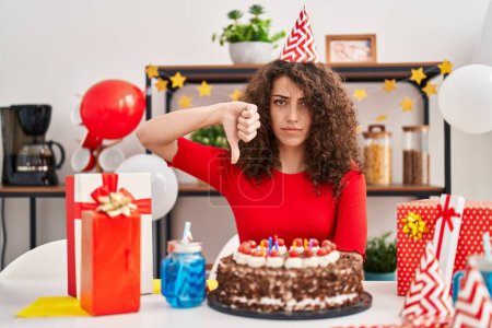 Téléchargez les photos : Femme hispanique avec les cheveux bouclés célébrant anniversaire tenant grand gâteau au chocolat avec le visage en colère, signe négatif montrant détester avec les pouces vers le bas, concept de rejet - en image libre de droit