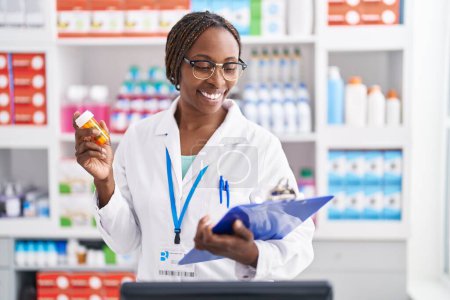 Photo for African american woman pharmacist holding pills bottle reading document at pharmacy - Royalty Free Image