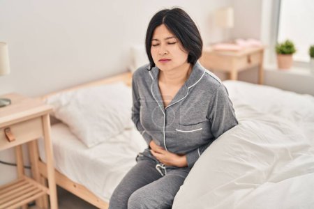 Téléchargez les photos : Jeune femme chinoise souffrant de douleurs menstruelles assise sur le lit dans la chambre - en image libre de droit
