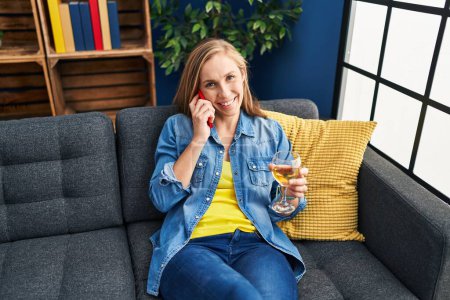 Téléchargez les photos : Jeune femme blonde parlant sur le smartphone et buvant du vin à la maison - en image libre de droit