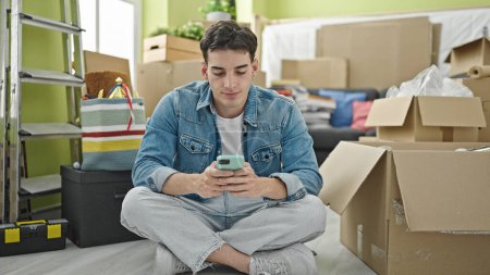 Foto de Hombre hispano joven usando teléfono inteligente sentado en el piso en casa nueva - Imagen libre de derechos