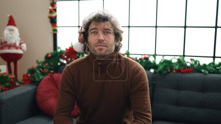 Téléchargez les photos : Jeune homme hispanique assis sur le canapé par la décoration de Noël avec une expression détendue à la maison - en image libre de droit