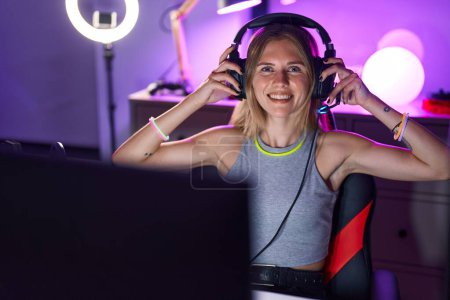 Téléchargez les photos : Jeune femme blonde streamer jouer à un jeu vidéo portant des écouteurs à la salle de jeux - en image libre de droit