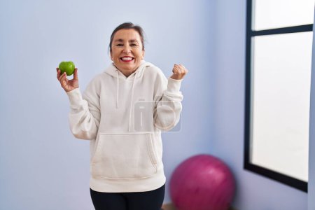 Photo for Middle age hispanic woman wearing sportswear holding healthy apple screaming proud, celebrating victory and success very excited with raised arm - Royalty Free Image