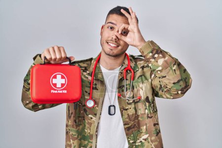 Foto de Joven médico hispano vistiendo uniforme del ejército de camuflaje sosteniendo botiquín de primeros auxilios sonriendo feliz haciendo buen signo con la mano en el ojo mirando a través de los dedos - Imagen libre de derechos