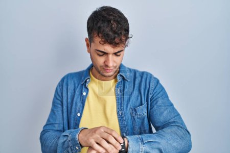 Photo for Young hispanic man standing over blue background checking the time on wrist watch, relaxed and confident - Royalty Free Image