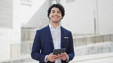 Téléchargez les photos : Jeune homme d'affaires latino-américain souriant confiant en utilisant le pavé tactile à la rue - en image libre de droit