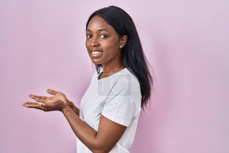 Téléchargez les photos : Jeune femme africaine portant casual t-shirt blanc pointant vers le côté avec les mains ouvertes paumes montrant l'espace de copie, présentant la publicité souriant heureux excité - en image libre de droit