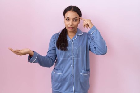 Téléchargez les photos : Jeune femme arabe portant un pyjama bleu confus et agacé avec la paume ouverte montrant l'espace de copie et pointant du doigt vers le front. Penses-y.. - en image libre de droit