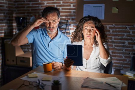 Photo for Middle age hispanic couple using touchpad sitting on the table at night worried and stressed about a problem with hand on forehead, nervous and anxious for crisis - Royalty Free Image