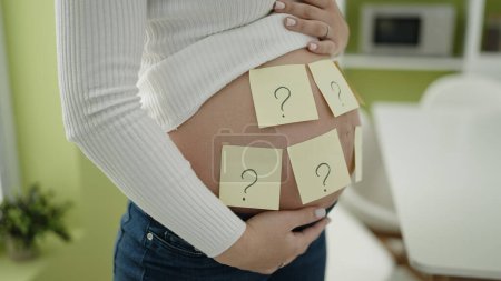 Téléchargez les photos : Jeune femme enceinte mettant papier rappel point d'interrogation sur le ventre demandant le sexe du bébé à la salle à manger - en image libre de droit