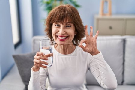 Foto de Mujer de mediana edad beber vaso de agua haciendo signo ok con los dedos, sonriendo gesto amistoso excelente símbolo - Imagen libre de derechos