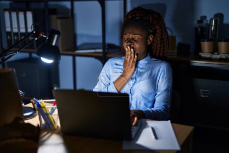 Foto de Mujer africana trabajando en la oficina por la noche aburrida bostezando cansada cubriendo la boca con la mano. inquietud y somnolencia. - Imagen libre de derechos