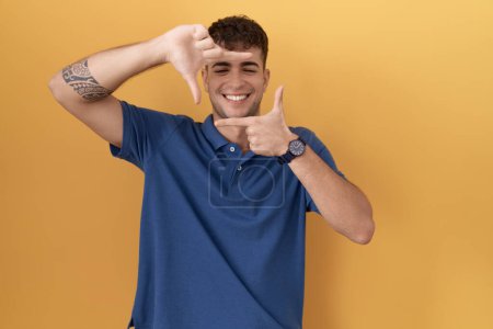 Téléchargez les photos : Jeune homme hispanique debout sur fond jaune souriant faisant cadre avec les mains et les doigts avec le visage heureux. concept de créativité et photographie. - en image libre de droit