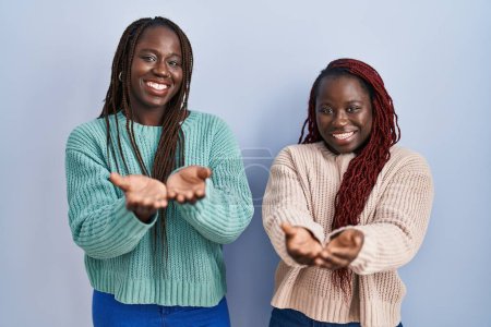 Foto de Dos mujeres africanas de pie sobre fondo azul sonriendo con las palmas de las manos juntas recibiendo o dando gesto. retención y protección - Imagen libre de derechos