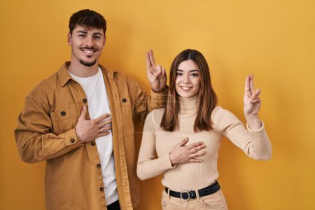 Photo for Young hispanic couple standing over yellow background smiling swearing with hand on chest and fingers up, making a loyalty promise oath - Royalty Free Image